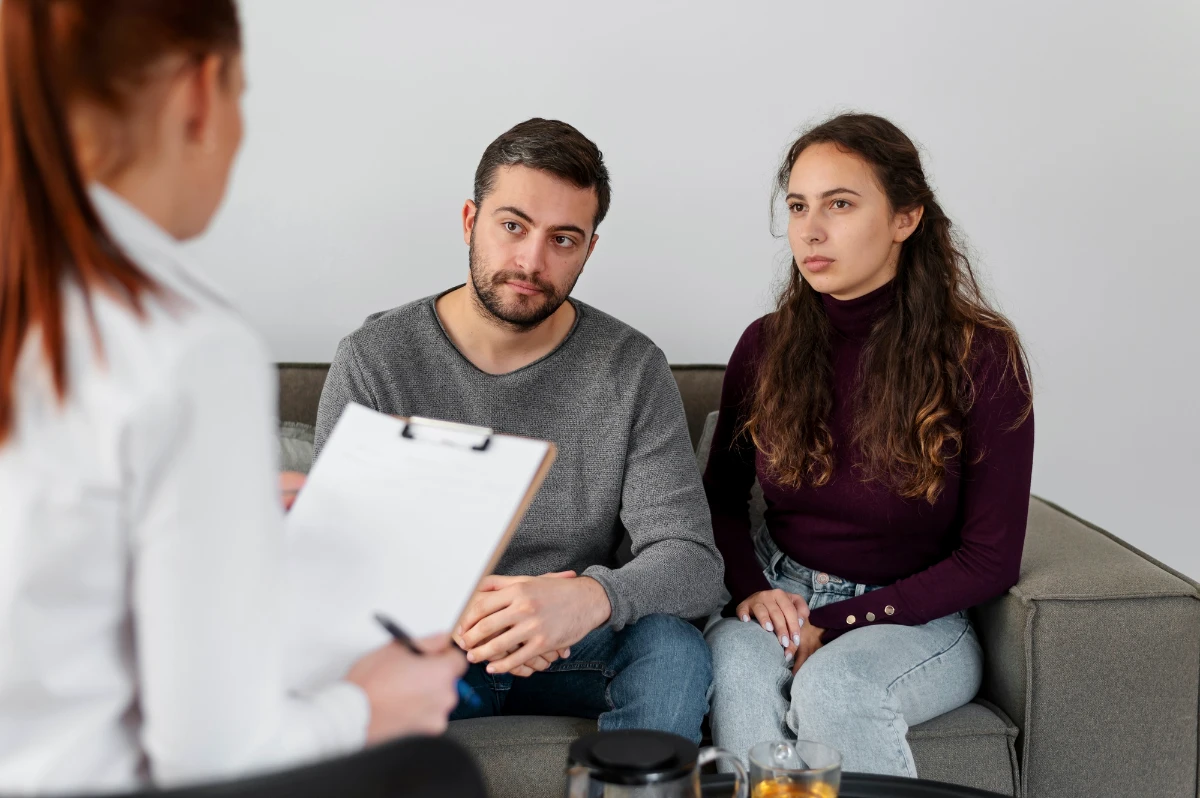A couple sitting together on a couch during a therapy session, listening attentively to a counselor taking notes.
